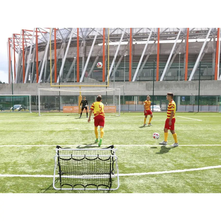 Fotball rebounder vegg, frontvisning på kunstgress med spillere i røde og gule drakter i bakgrunnen.
