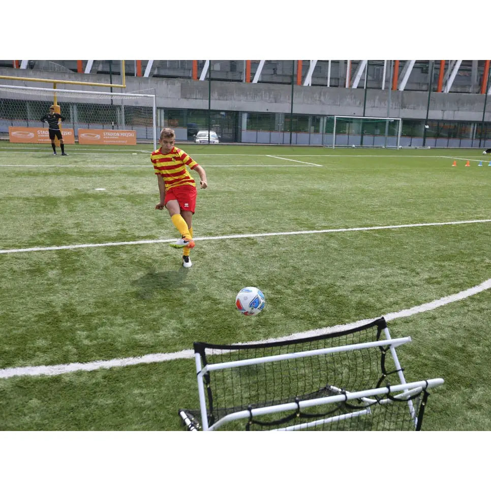 Fotball rebounder nett, sett bakfra mens en gutt i rød og gul drakt skyter ball mot nettet på kunstgress.