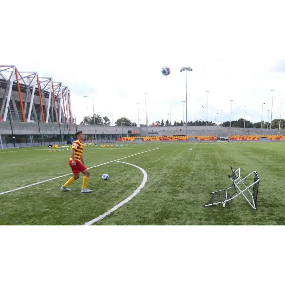 Fotball rebounder vegg på kunstgress, spiller i aksjon med ballen i lufta foran sort nett.