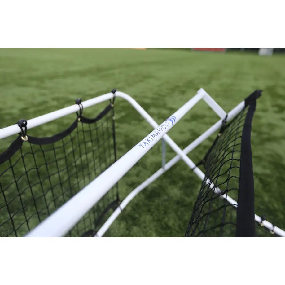 Fotball rebounder vegg med hvit ramme og sort nett, sett skrått ovenfra på kunstgress med synlig logo.