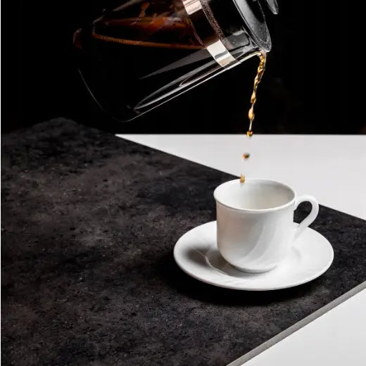 Hvit porselenskopp med skål, kaffe helles fra presskanne, plassert på mørk overflate mot lys bakgrunn.