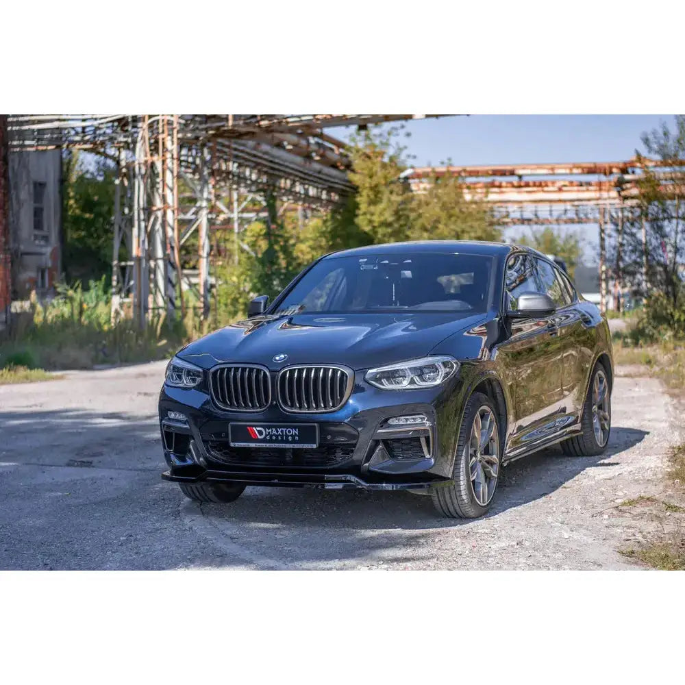 Frontleppe BMW X4 M-Pack G02 med sorte detaljer, sett skrått forfra på grusvei foran industribygg.