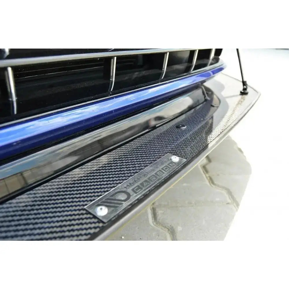 Front lip på svart karbonfiber spoiler til VW Golf VII R med metallskilt og synlig logo, blå støtfanger over.