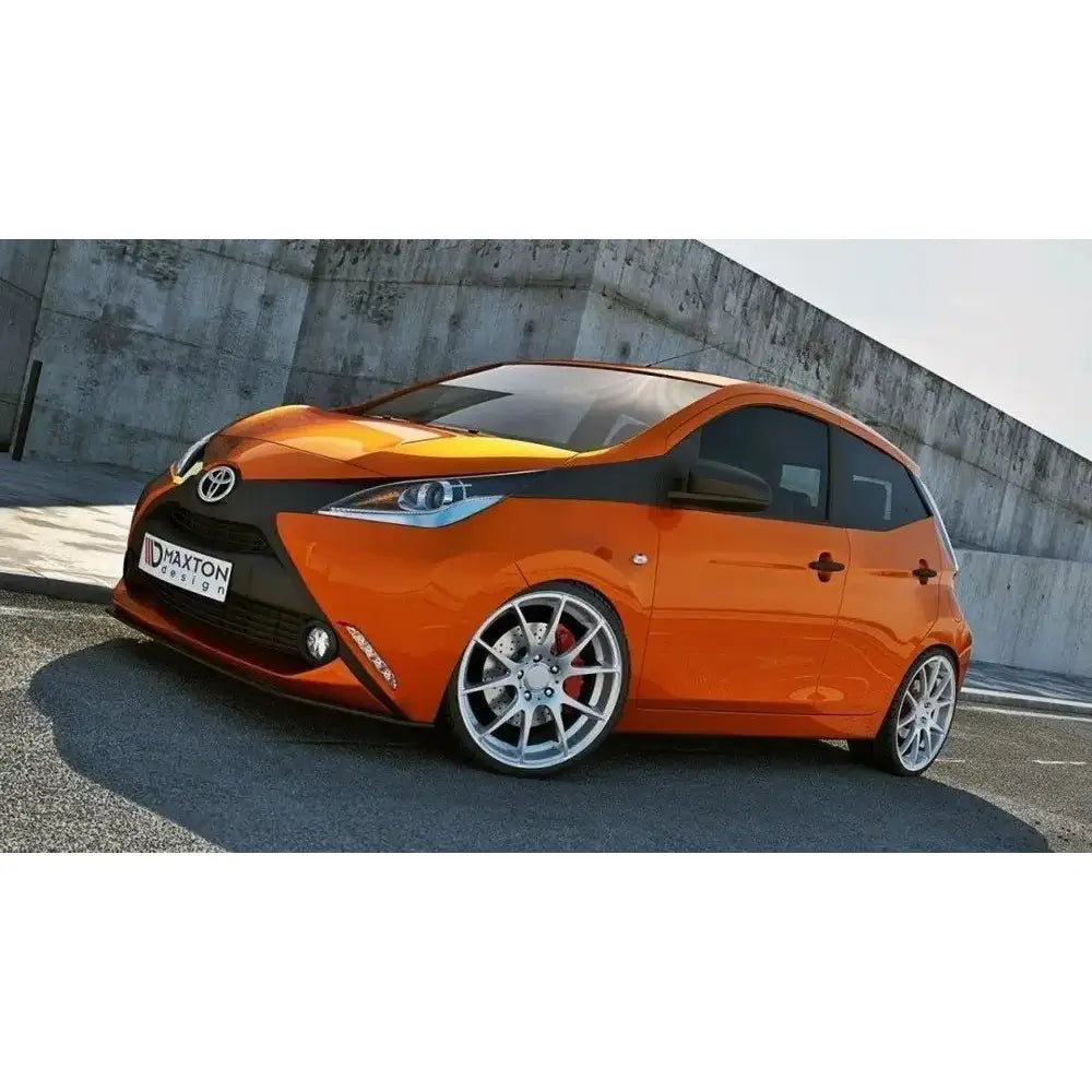 Frontleppe på Toyota Aygo Mk2, oransje bil med svarte detaljer og sølvfelger ved betongvegg.