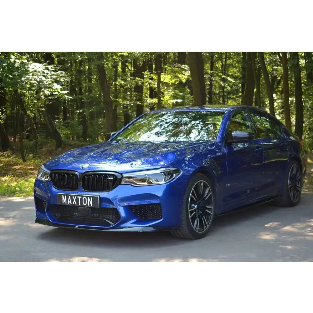 Front lip på BMW M5 F90 V.1, blå sportsedan med sorte felger sett skrått forfra på vei i skog.