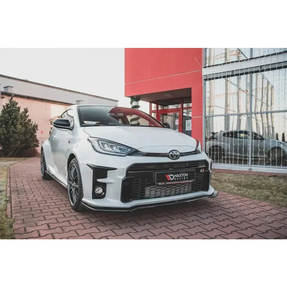 Front lip på Toyota GR Yaris Mk4 V.1, hvit bil med svart frontspoiler og aluminiumsfelger parkert ute.