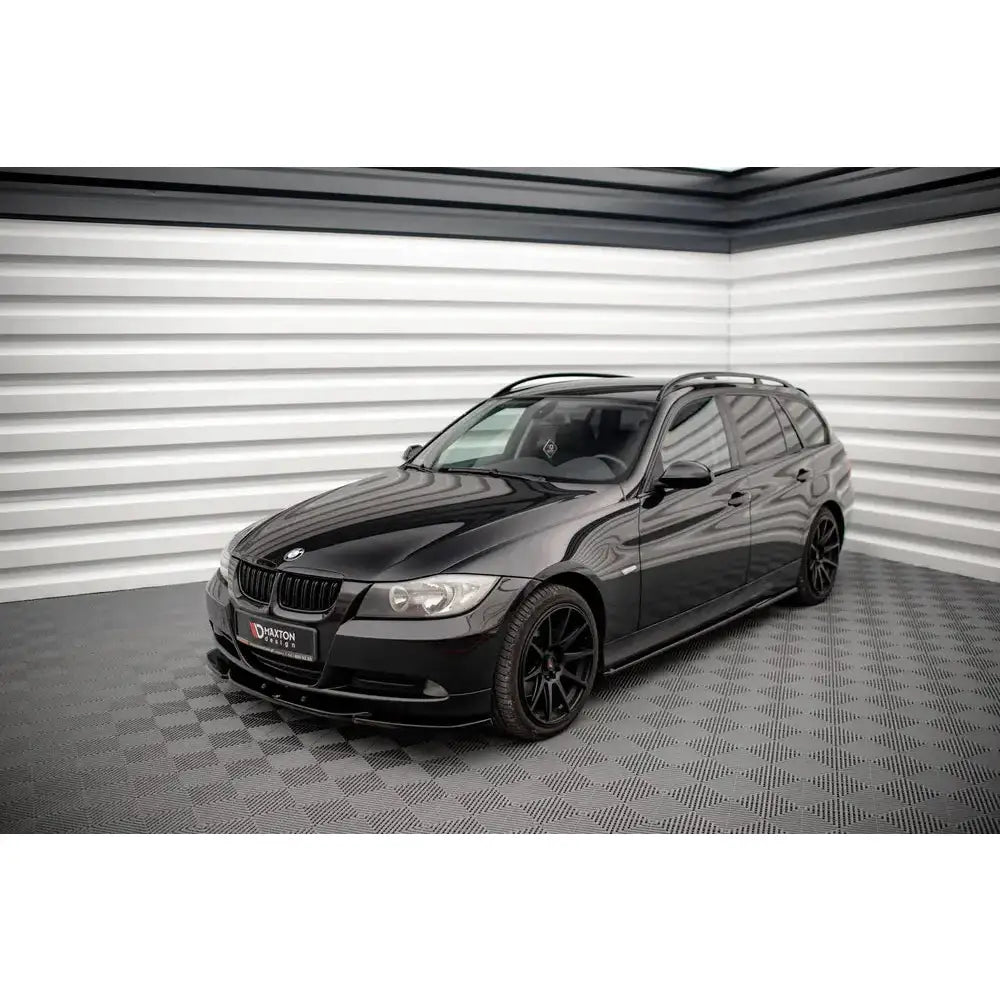 Front splitter på svart BMW 3 E90 stasjonsvogn med svarte felger og takrails, fotografert innendørs.