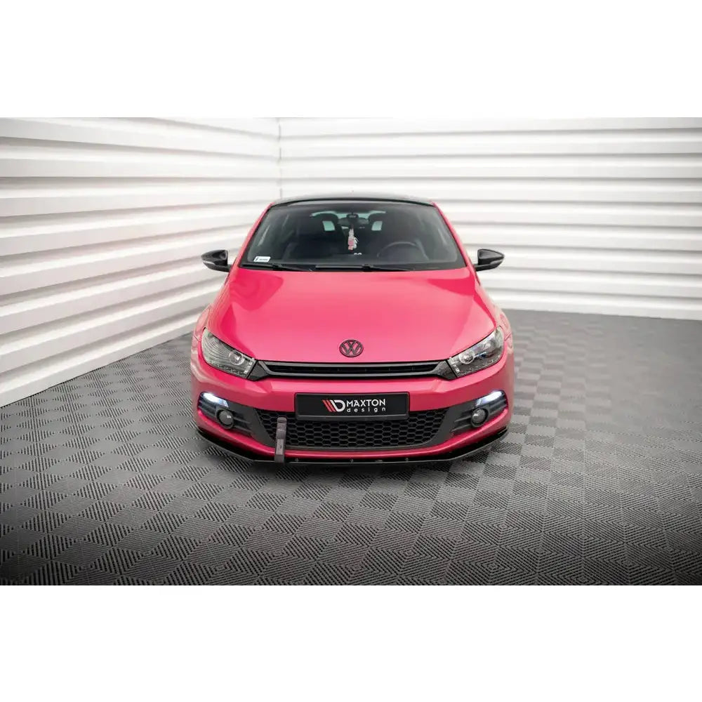 Spoiler på VW Scirocco, rød bil sett forfra med mørk grill og synlig logo mot hvit riflet vegg.