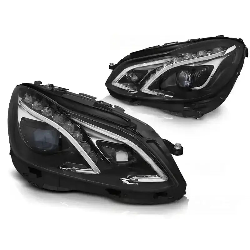 Hovedlykter Mercedes W212 13-16 med sotet glass og LED-striper, vist fra skrå vinkel mot hvit bakgrunn.