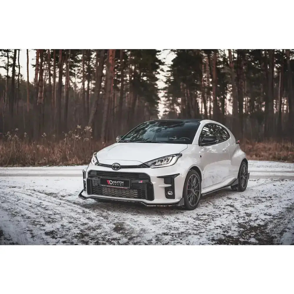 Frontleppe på hvit Toyota GR Yaris Mk4 med svarte felger, parkert på snødekt grusvei foran skog.