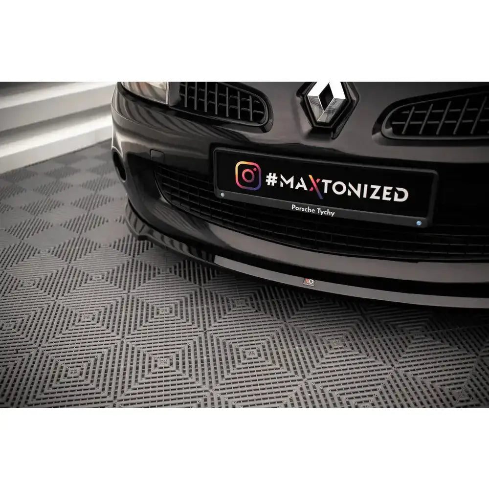 Front splitter for Renault Clio III Rs på svart bil med dekorativt skilt, nærbilde på grått gulv.