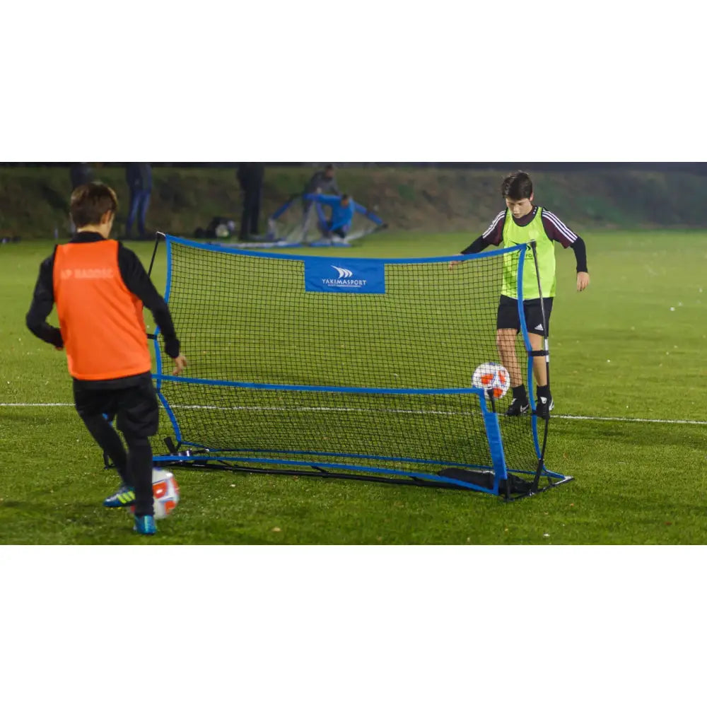 Galaktisk Hoppeseng med blå ramme, barn spiller fotball over nettet på grønn gressbane under utendørstrening.