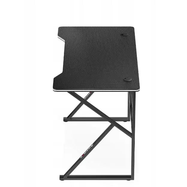 Gamingbord Huzaro Hero 1.6, toppvisning av svart karbonfiberlignende bordflate med kabelhull og metallben i kryss.