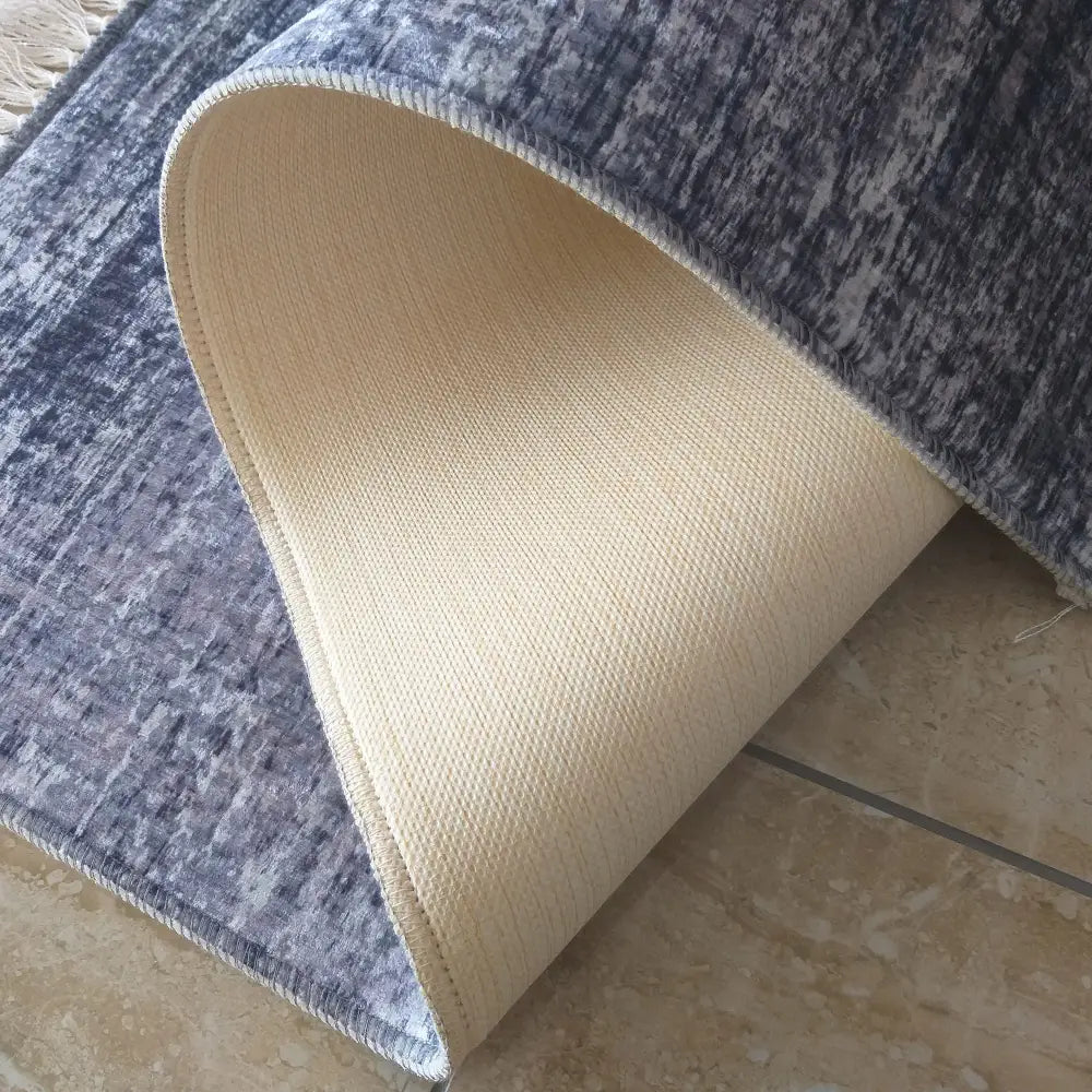 Grå og beige teppe 160 x 220 cm, nærbilde av oppbrettet hjørne som viser sklisikker bakside og tekstur.