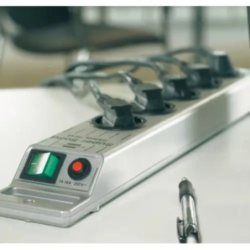 Grenuttak med overspenningsvern i sølv, fem uttak og grønn bryter, sett skrått på bord med tilkoblede kabler.