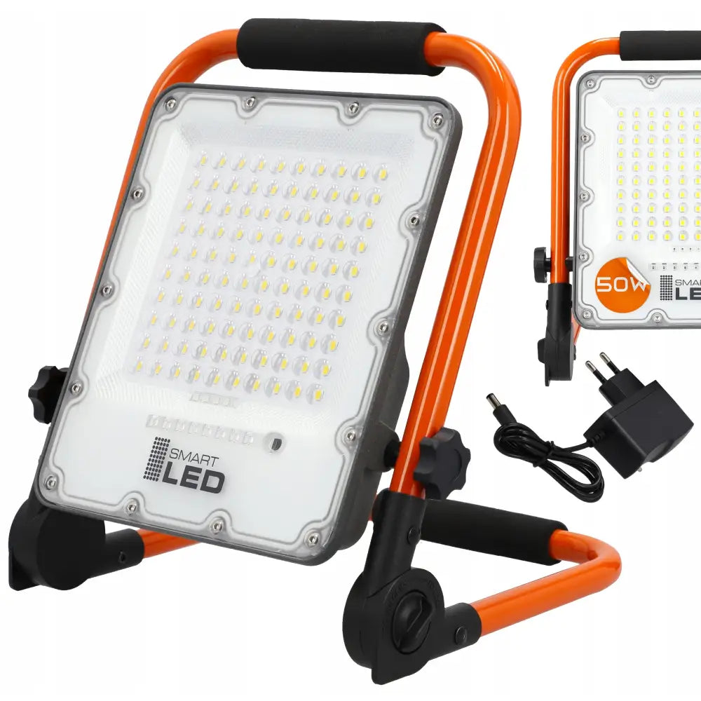 LED byggelyskaster 50W på oransje stativ, sett skrått forfra med lader og støpsel ved siden av.