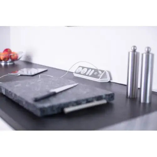 Sort skjærefjøl i stein med kniv oppå, plassert på kjøkkenbenk foran stålkverner og eplebolle.