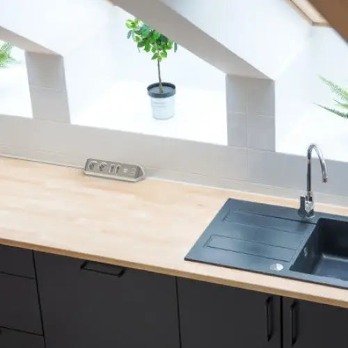 Hjørneuttak estilo V2 med fire stikk og to USB, montert på kjøkkenbenk ved sort vask og krom kran.