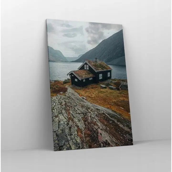 Lerretsbilde med naturmotiv, hytte ved innsjø og fjell, sett skrått mot hvit vegg for moderne interiør.