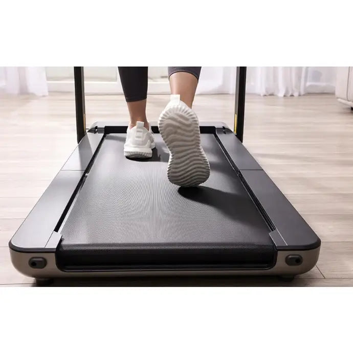 Tredemølle Kingsmith Walkingpad X21, bakfra med bruker i hvite joggesko på lyst tregulv og gardiner.