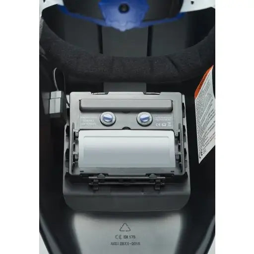 Innsiden av hjelm for invertersveisemaskin, nærbilde av batterirom med to batterier og varsellapp.