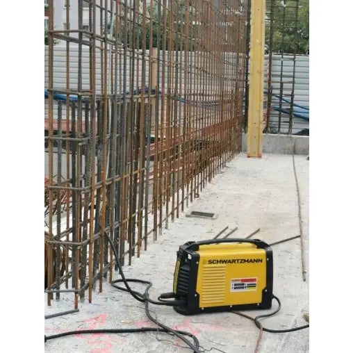 Invertersveisemaskin Tig Xxl Sett Schwartzmann på byggeplass, omgitt av armeringsjern og betonggulv, tilkoblet ledninger.