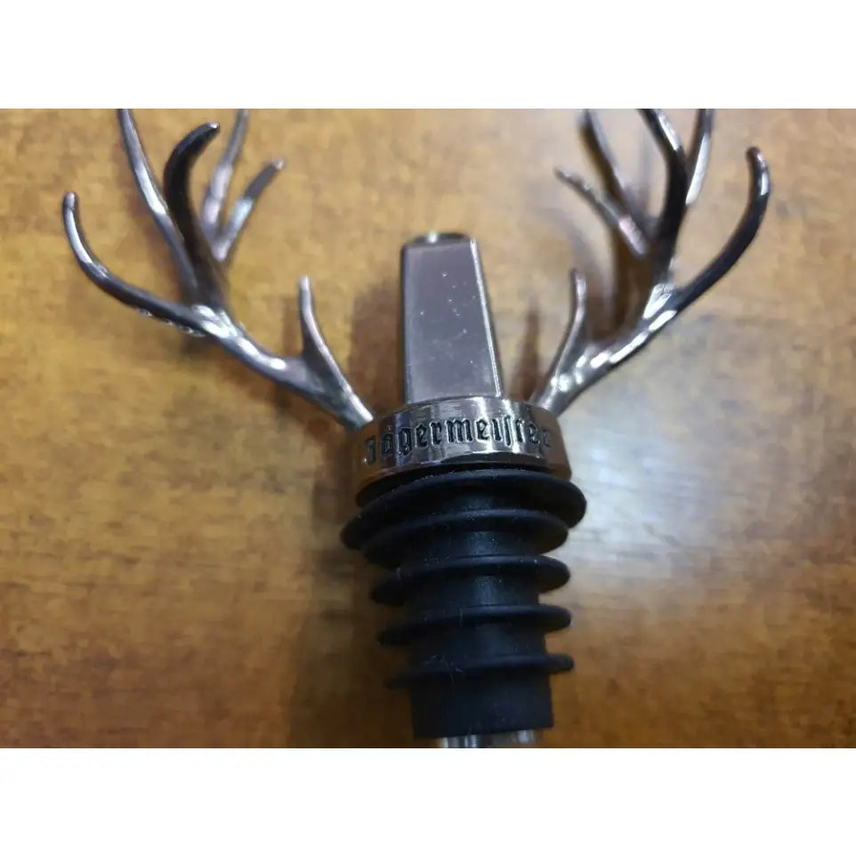 Jägermeister tappeapparat med sølvfargede hjortehorn og inngravert metallpropp, nærbilde mot brun bakgrunn.