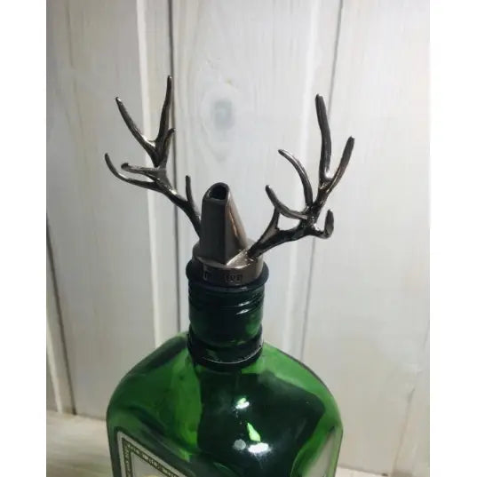 Jägermeister tappeapparat med hjortehornformet metalltut, grønn flaske mot lys trevegg, skrå vinkel.