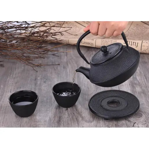 Jern tepotte sett med sort tekanne som heller te i kopp, to kopper og underlag på rustikk trebord.