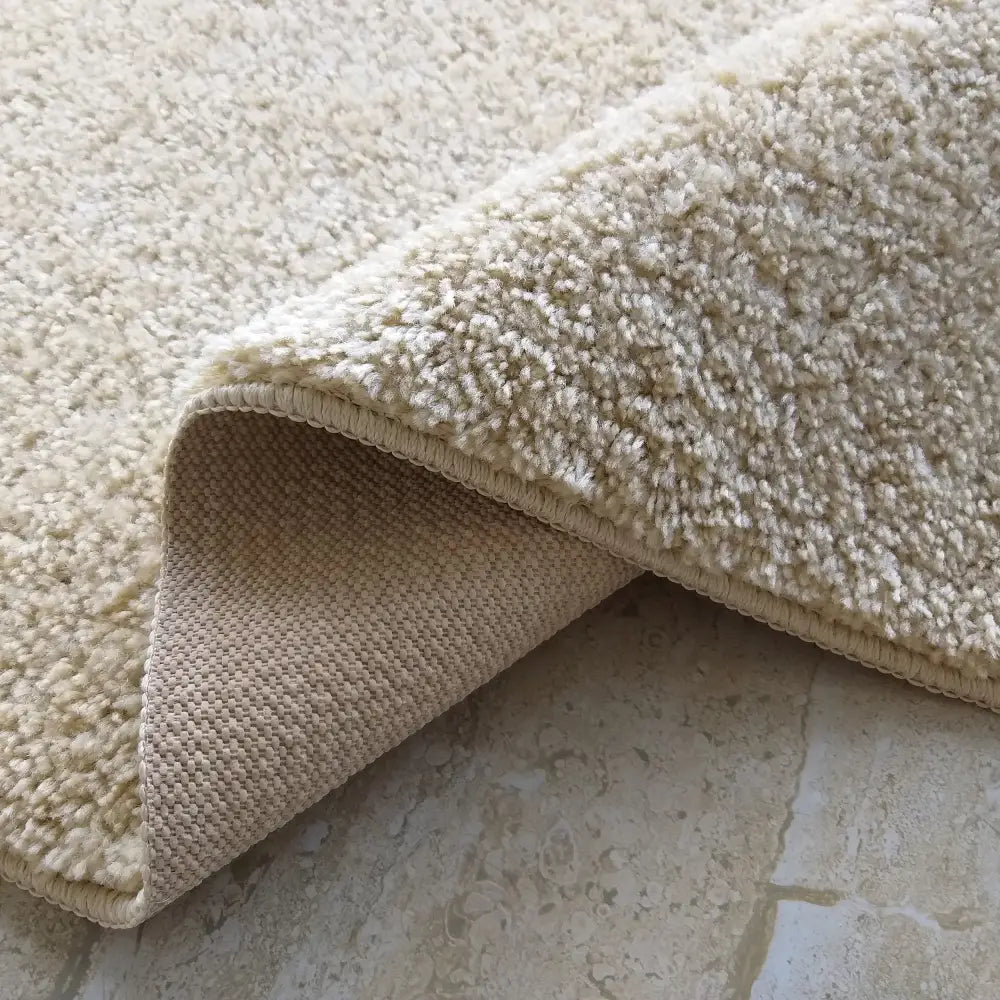 Beige teppe, nærbilde med oppbrettet hjørne som viser myk overflate og teksturert bakside.