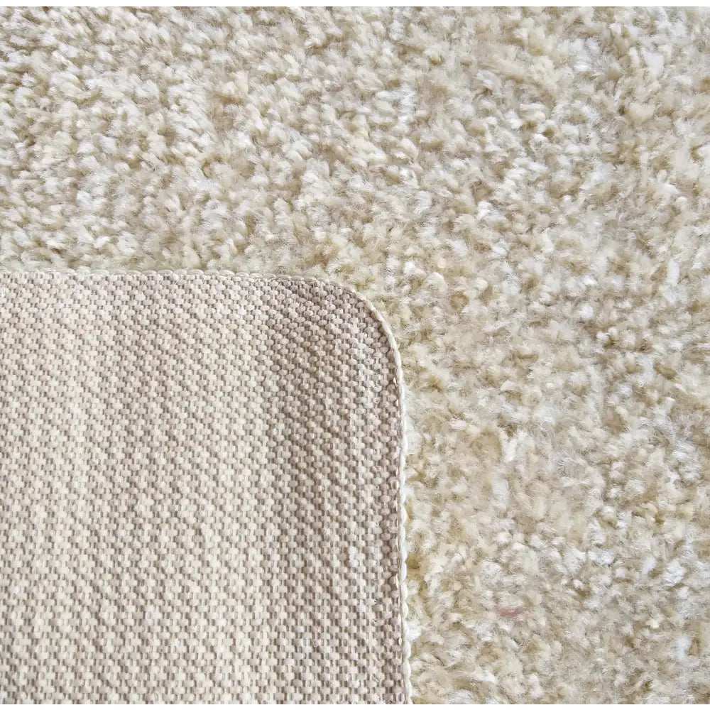 Kamel teppe 200 x 290 cm, nærbilde av undersidens vevde struktur og myk, lys beige lugg med avrundet hjørne.