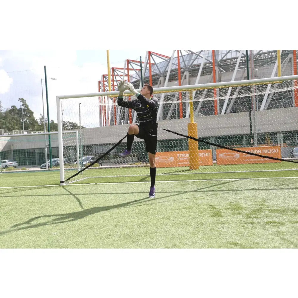 Keeper Treningkummi, fotballkeeper i svarte klær hopper sideveis foran mål for å fange ball på grønn bane.