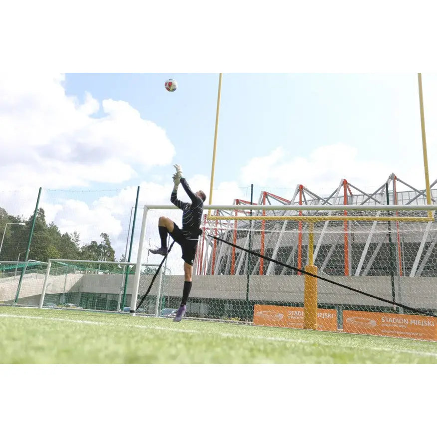 Keeper Treningkummi, fotballkeeper i svart drakt hopper for å redde ball foran mål på gressbane med tribune.