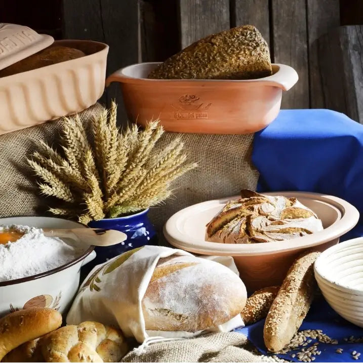 Keramisk brødform med lokk, fylt med hjemmebakt brød, omgitt av ulike brød, korn og bakeutstyr på blå duk.