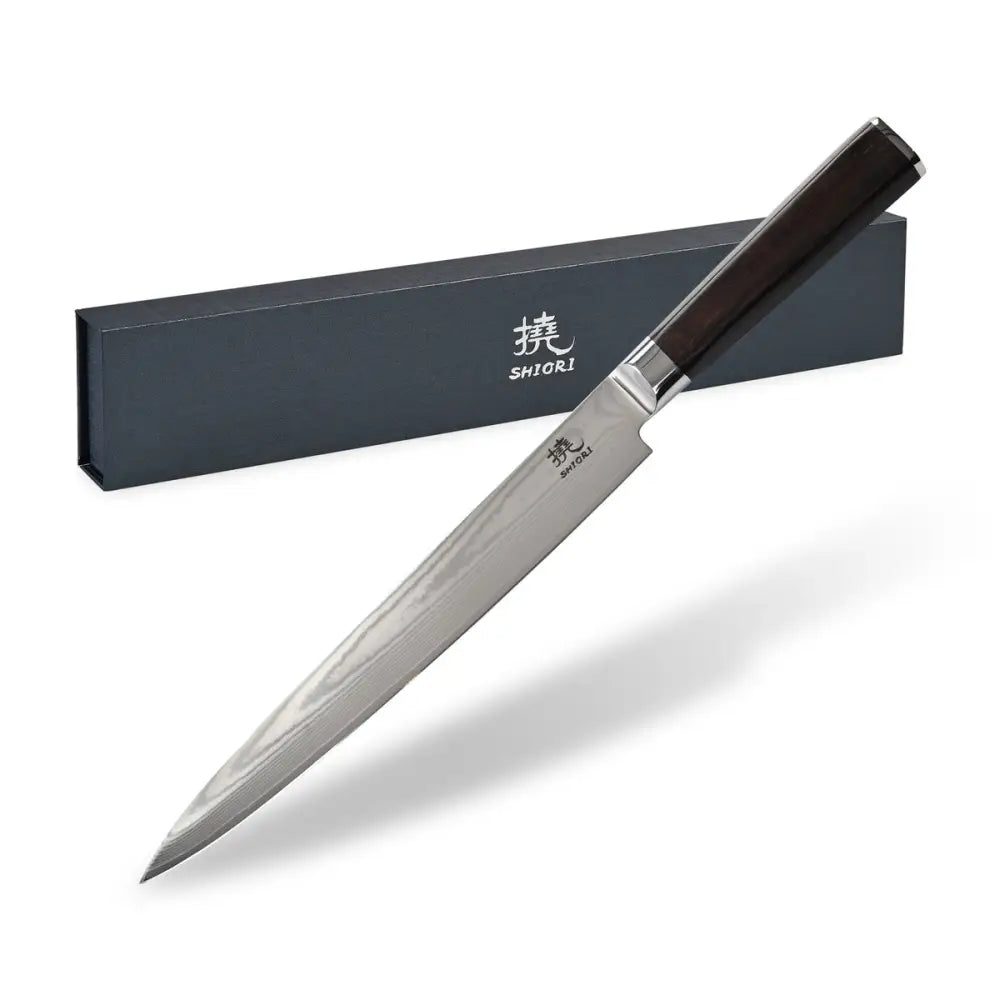 Kjøkkenkniv i japansk damaskstål med mørkt ebony-håndtak, skråvisning foran sort gaveeske med logo.
