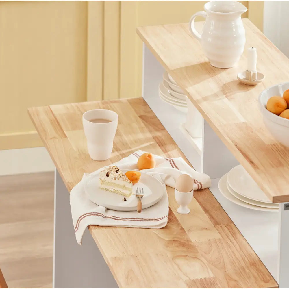 Kjøkkenøy med skap, lyst trebord sett ovenfra med kakestykke, aprikoser og servise på bordplaten.