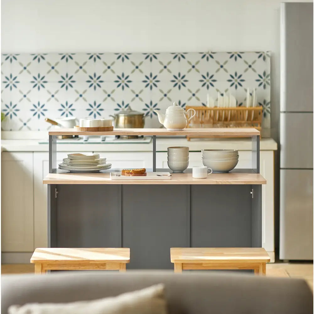 Kjøkkenøy med lyst treverk, mørkegrå base og to barkrakker, dekket med servise og bakverk mot flisvegg.