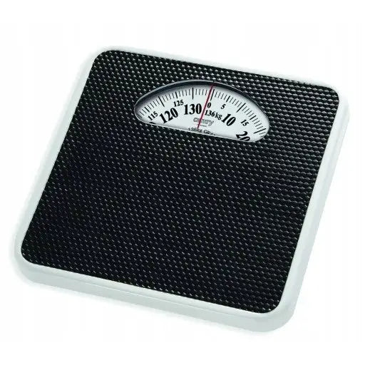 Klassisk svart mekanisk badevekt med hvit ramme, skrått ovenfra, tydelig analogt display og mønstret overflate.