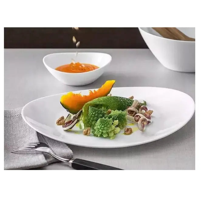 Komplett servise sett, hvit oval middagstallerken med grønnsaker, nøtter og oransje saus på grå duk.