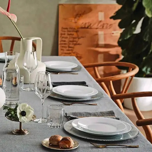 Komplett servise sett Toledo, hvite tallerkener og vinglass på grå duk, sett skrått med trestoler rundt bordet.