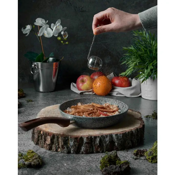 Grå Konighoffer stekepanne sett fra siden med mat, krydder drysses over, frukt og grønne planter i bakgrunnen.
