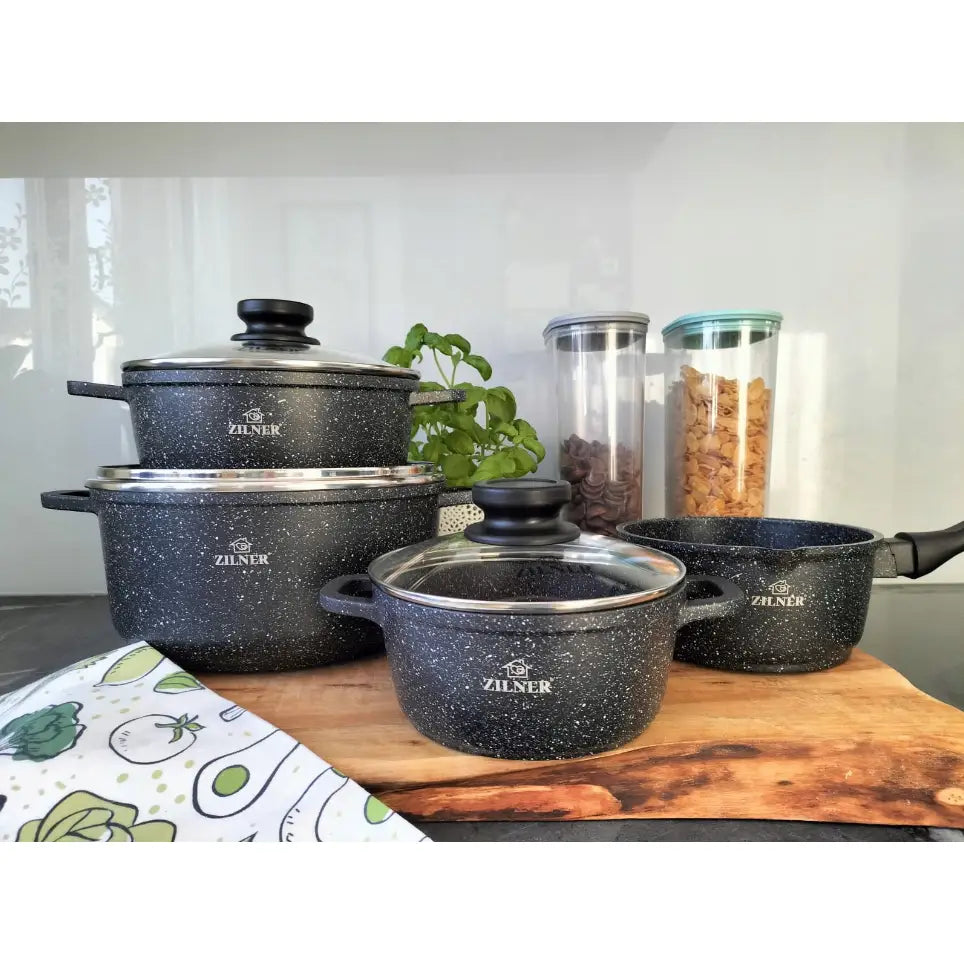 Komplett sett med sorte, granittmønstrede gryter og kjeler med glasslokk og håndtak på trefjøl på kjøkkenbenk.