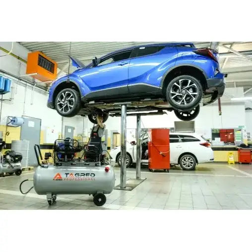 Kompressor 100L med tre stempler, plassert på verkstedgulv foran løftet blå bil og verktøyskap.