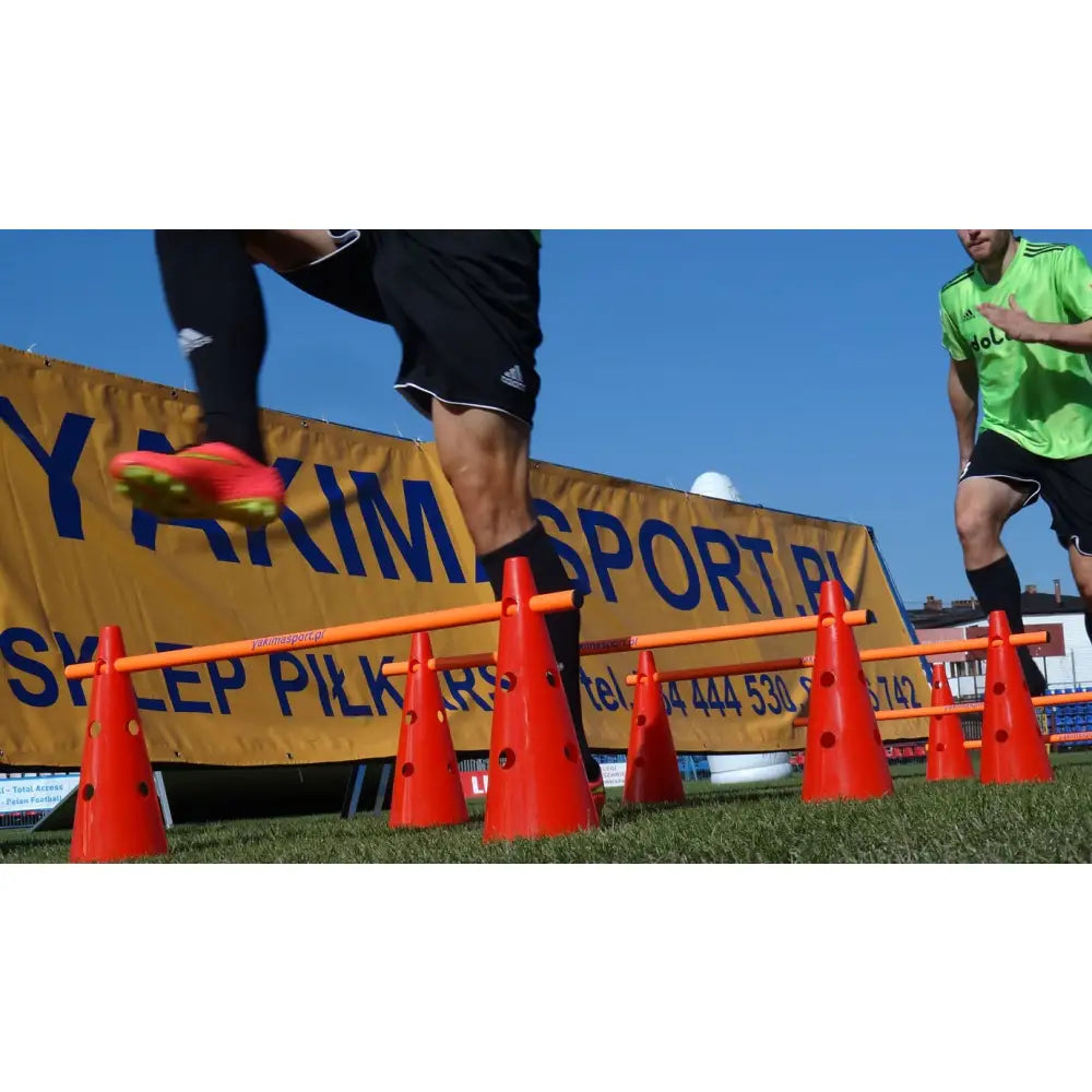 Koordineringssett med to oransje kjegler og stang, brukt som hinder mens to personer hopper utendørs på gress.