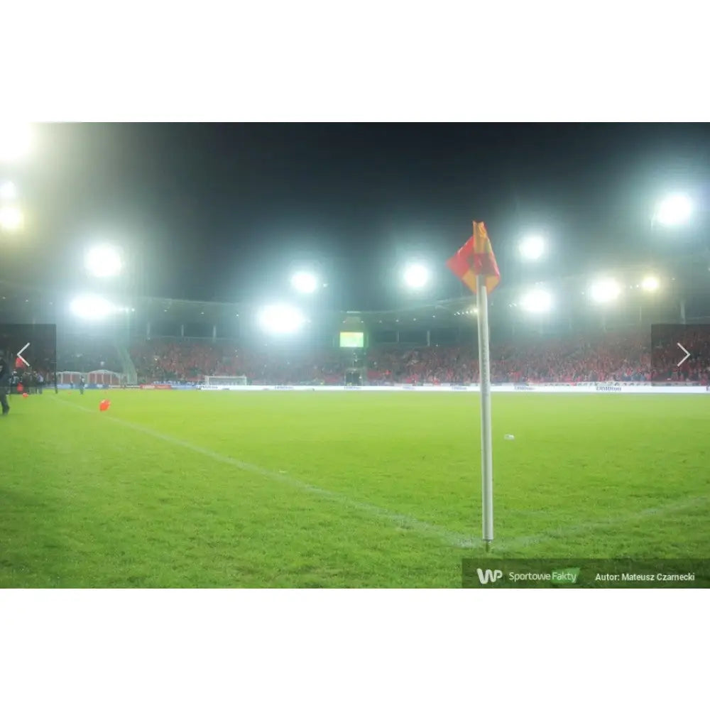 Kornerflagg med rød-gul vippbar stang plassert i hjørnet av grønn fotballbane, stadionlys bak.