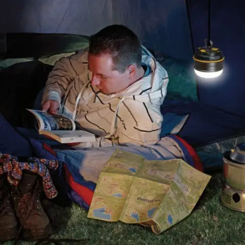 Lampe LED OLI, rund lykt henger i telt og lyser mens mann leser i sovepose med turutstyr rundt.