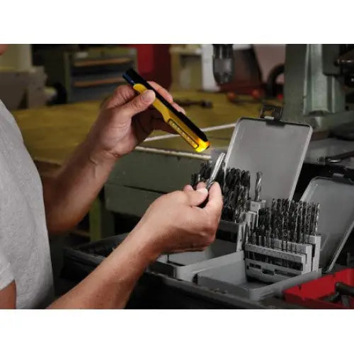 LED PEN LIGHT, gul og svart, holdes i hånd sammen med drillbit foran åpen borboks på verkstedbenk.