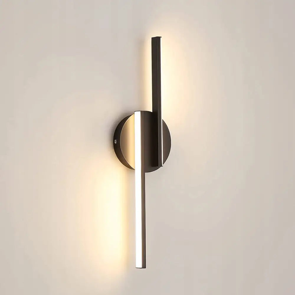 LED vegglampe med sort rund base og to opplyste vertikale staver i matt metall mot lys vegg.