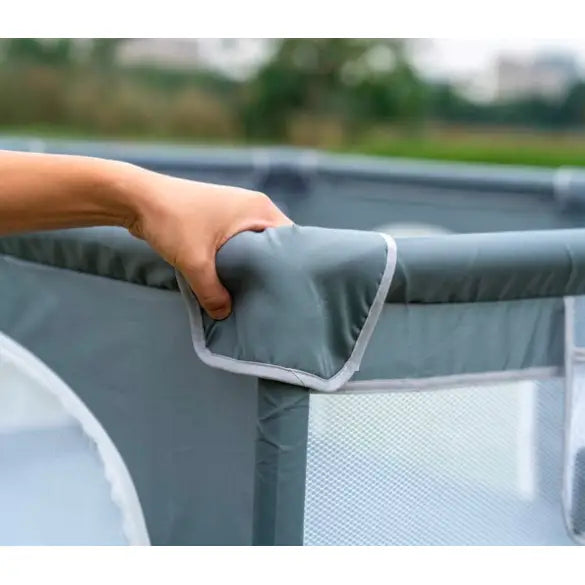 Lekegrind for barn med grått stoffgjerde, hvite kanter og polstret hjørne holdt av en hånd, nærbilde utendørs.