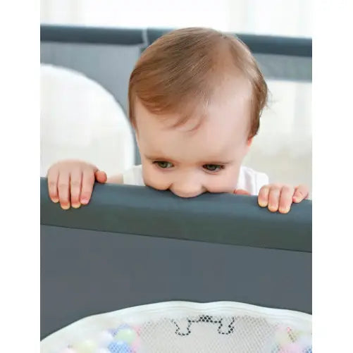 Lekegrind for barn med mørkgrå ramme, baby med lyst hår biter forsiktig i kanten, nærbilde forfra.